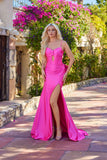A woman with long blonde hair wears the Juliet JT2728H long prom dress in fuchsia, featuring a thigh-high slit, as she stands on a stone path lined with flowers and greenery.