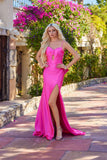 A woman with long blonde hair stands outdoors in the Juliet JT2728H long prom dress—bright pink, tight-fitting with thin straps, a front cutout, and a thigh-high slit—amid blooming flowers and stone walls.