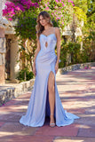 A woman stands outdoors on a stone walkway, surrounded by greenery and purple flowers, wearing the Juliet JT2728H Long Prom Dress in periwinkle—a fitted gown with spaghetti straps and a thigh-high slit