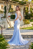 A woman wearing the Juliet JT2727H Long Prom Dress—a fitted, light blue jersey gown with a lace-up back and floor-length train—stands outdoors on a stone pathway, glancing over her shoulder.