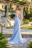 A woman wearing the Juliet JT2727H Long Prom Dress—a fitted, light blue jersey gown with a lace-up back and floor-length train—stands outdoors on a stone pathway, glancing over her shoulder.