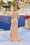 A woman stands by a blue-tiled pool wearing the Juliet JT2723C long prom dress, featuring a fitted, floor-length silhouette with a one-shoulder neckline, intricate beading, and sheer panels, with flowers and white balustrades behind her.