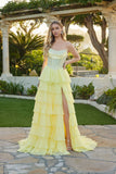 A woman stands outdoors by greenery and stone architecture, wearing the Juliet JT2738S Long Prom Dress in yellow, featuring a strapless design, tiered chiffon ruffle skirt, and high slit.