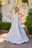A woman stands outside on a tiled walkway, wearing the Juliet JT2742A long prom dress featuring a light floral texture, fitted corset bodice, and full skirt, with a staircase and greenery in the background.