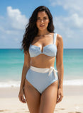 Woman in a light blue bikini standing on a beach with clear blue water and sky.